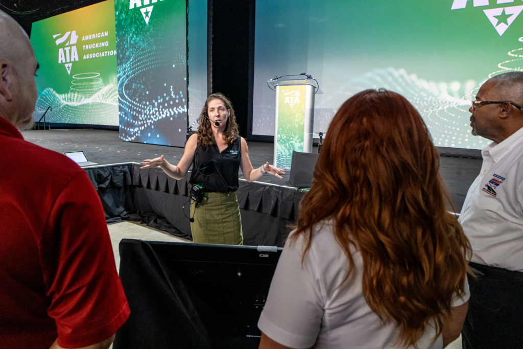 A Woman Wearing A Headset Microphone Stands In Front Of A Stage, Gesturing With Both Hands As She Speaks To Three People. The Stage Backdrop Displays The American Trucking Association ( A T A) Logo With Green And Blue Digital Wave Graphics. The Woman Appears To Be Giving Instructions Or Leading A Briefing, Possibly For An Event Or Presentation Setup. One Man Wears A Red Shirt, A Woman With Long Red Hair Wears A White Shirt, And Another Man Wears A White Shirt With A Logo On The Sleeve.