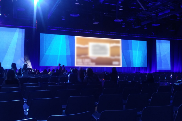 Large conference room with a stage featuring an expansive multi-screen display with blue geometric visuals. Audience members are seated and conversing as they wait for the presentation to begin.